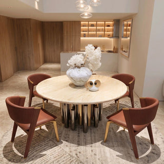 A round travertine dining table with a light wood top and metallic cylindrical base, surrounded by four brown upholstered chairs with gold-toned legs, set on a patterned rug in a modern dining room.